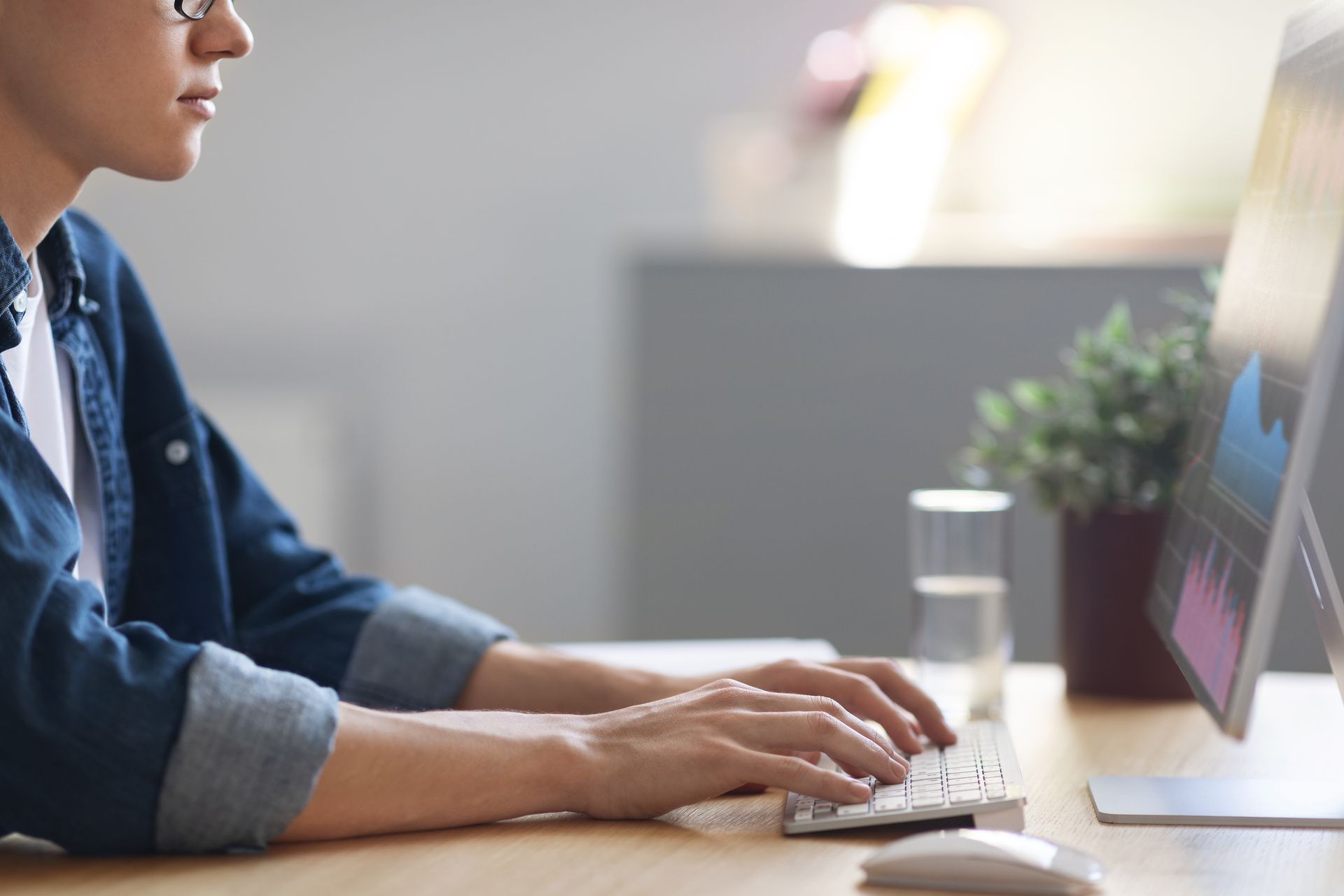 Person typing on a keyboard at a desk, looking at a computer monitor with graphs, plant and glass of water.