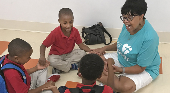 Teacher and three students seated on the floor, holding hands. Teacher wears glasses and a teal shirt.