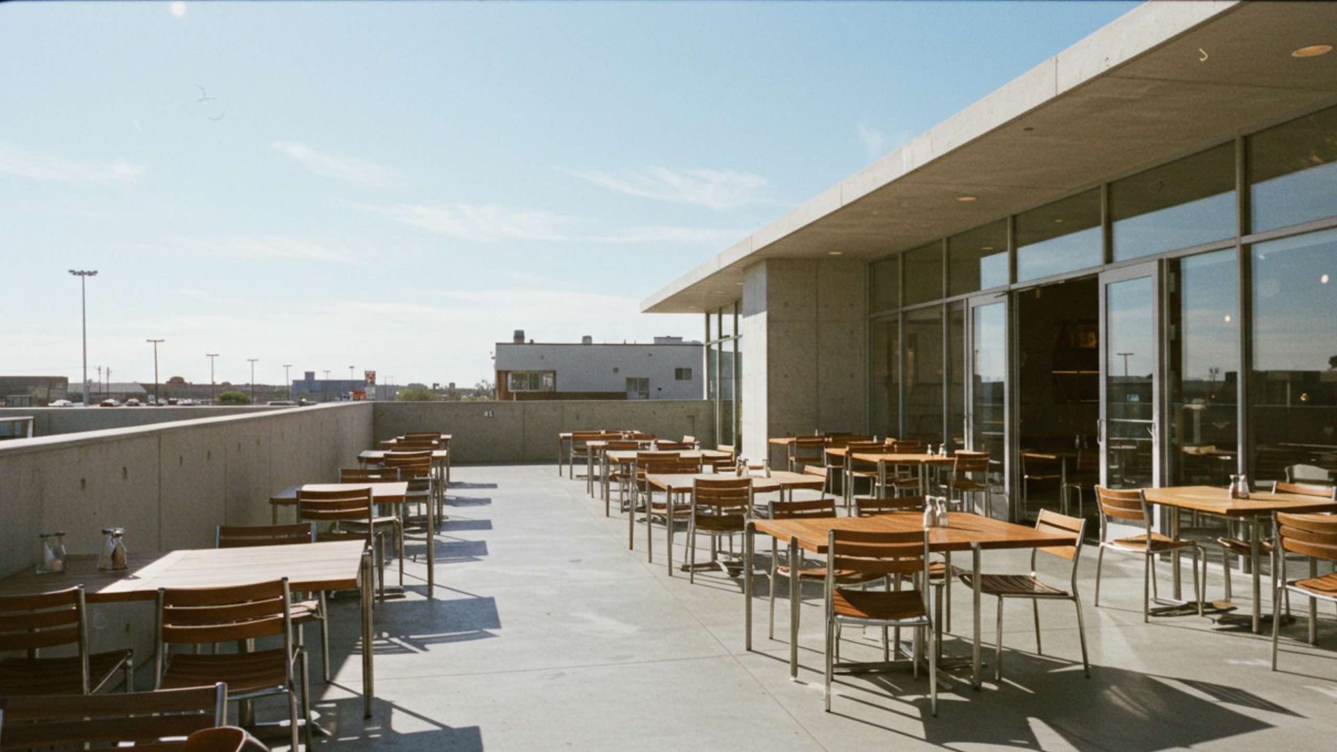 Outdoor cafe with wooden tables and chairs on a concrete patio next to a building with large windows.