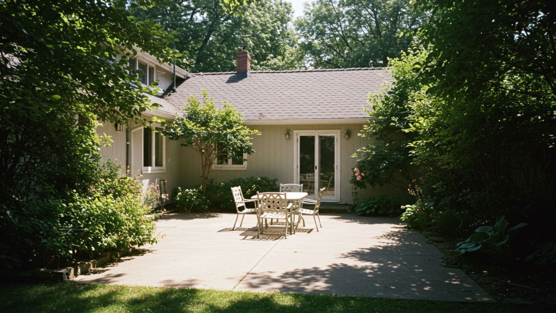 Patio with table and chairs in front of a house, surrounded by trees.