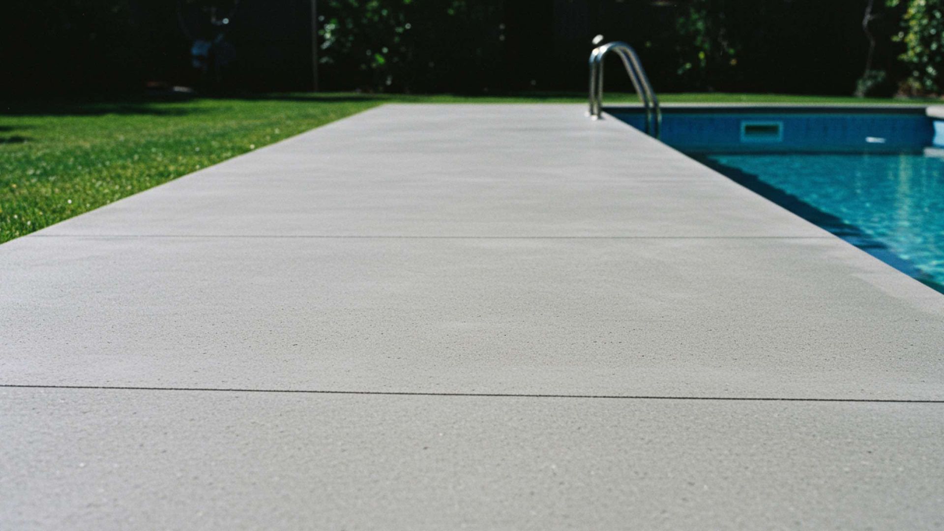 A smooth gray concrete pool deck extends alongside a rectangular blue swimming pool with metal ladders.