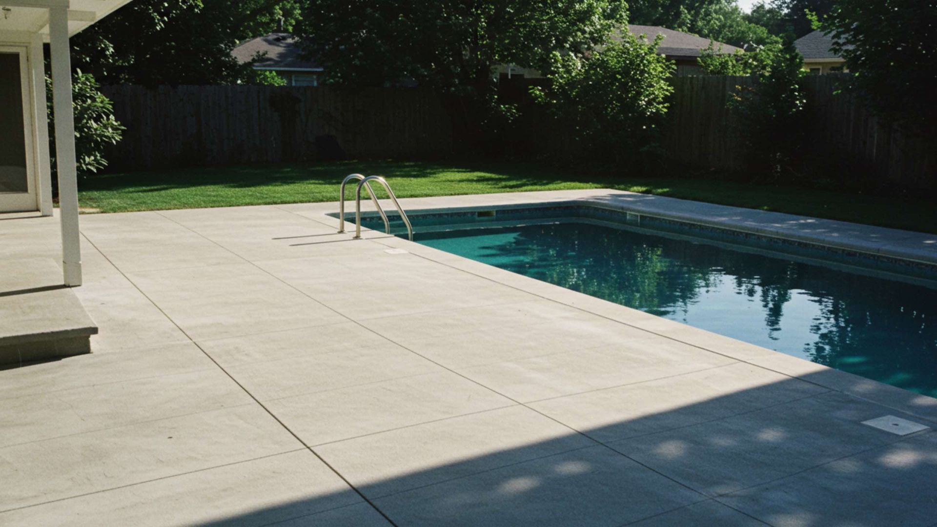 A rectangular swimming pool in a residential backyard, bordered by a large concrete deck under bright sunlight.