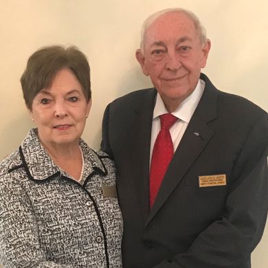 Two people standing together indoors, one in a black suit with a red tie and one in a patterned jacket.