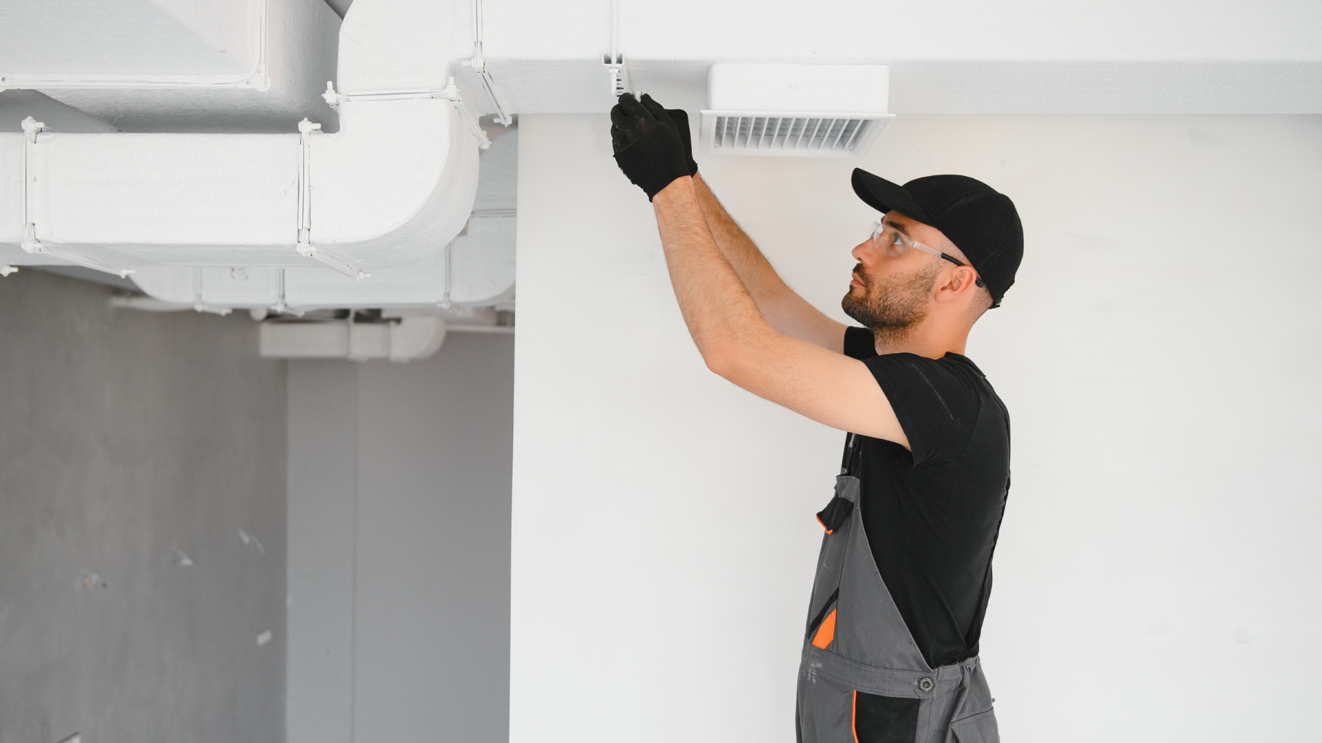 Man in work clothes installing an air vent on a white wall; ducts visible.