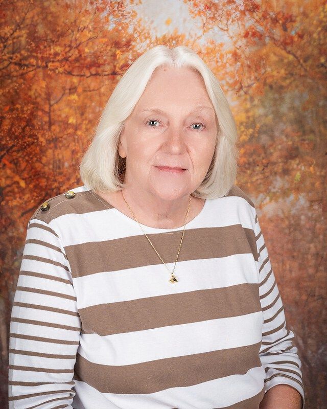 Portrait of a woman with white hair wearing a brown-and-white striped shirt against an autumn backdrop