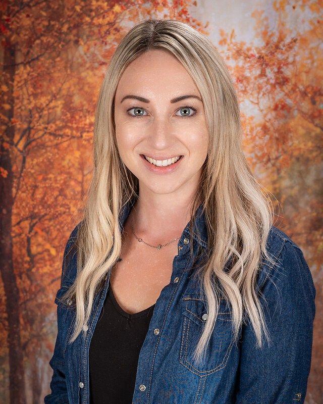 Smiling woman in a denim shirt posed against an autumn foliage backdrop