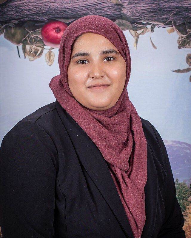 Mrs. Tauheed Ali - A woman in a maroon hijab and black blazer, smiling against a soft floral backdrop