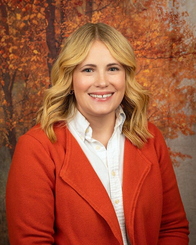 Smiling woman in an orange blazer and white shirt, posed against a warm autumn leaf backdrop