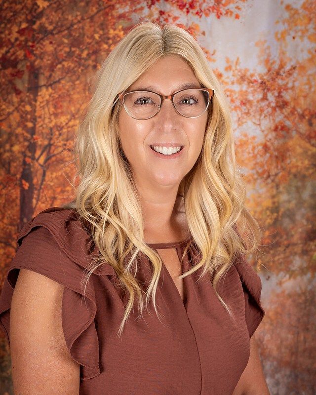 Smiling person with glasses and blonde hair in a brown top against an autumn leaves backdrop