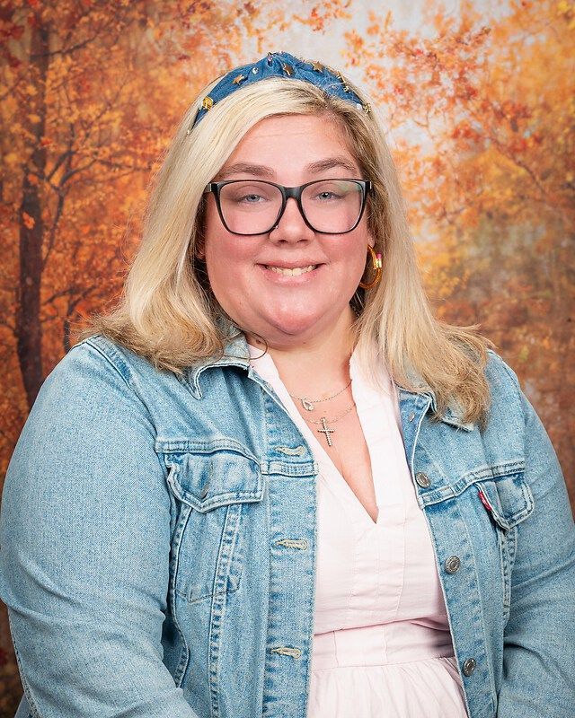 Smiling person with glasses in denim jacket against an autumn backdrop