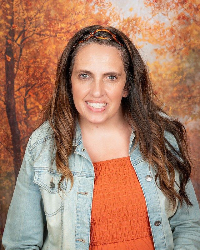 Woman smiling in a denim jacket and orange top against an autumn forest backdrop