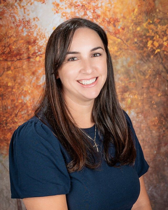 Smiling woman with long dark hair in a navy shirt against a warm orange studio backdrop