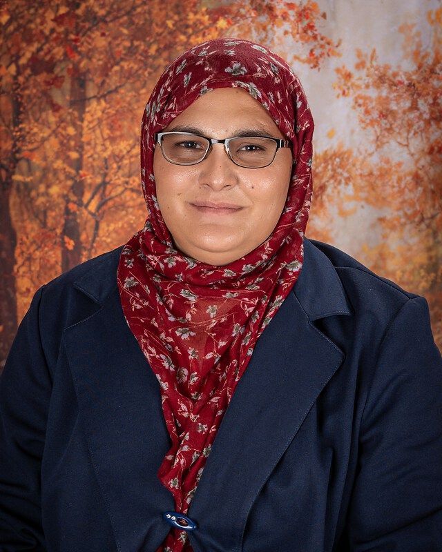 Portrait of a smiling person in glasses, red patterned hijab, and navy blazer against an autumn backdrop