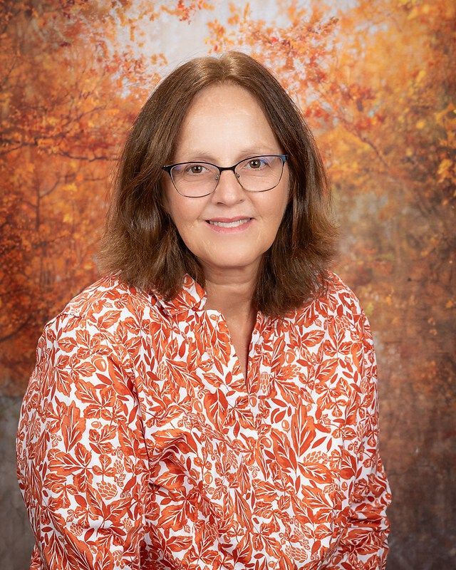 Portrait of a smiling woman in glasses wearing an orange patterned blouse against an autumn backdrop
