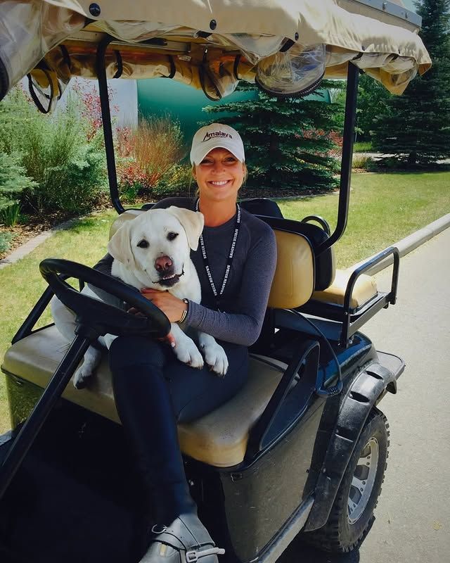 Woman and yellow Labrador dog sit in golf cart. Dog rests on lap and smiles. Outdoors with trees.