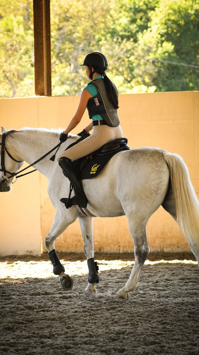 Woman riding a gray horse in an arena; wearing helmet, vest, and boots.