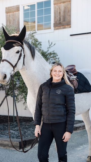 Smiling woman standing beside a saddled black-and-white horse outdoors by a white building