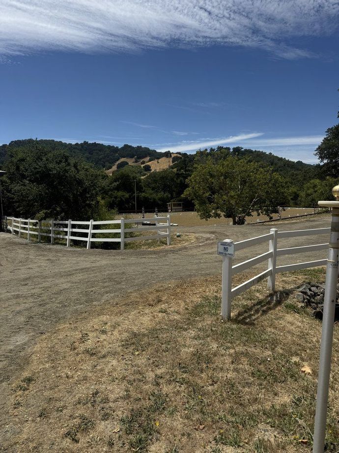 White fence and gravel driveway leading towards trees and hills under a blue sky.