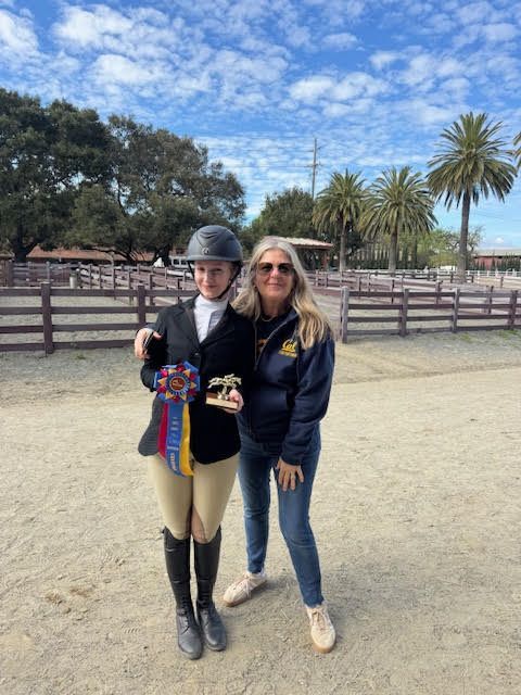 Two women standing together outdoors, one in a riding helmet holding colorful ribbons, near a fenced field.