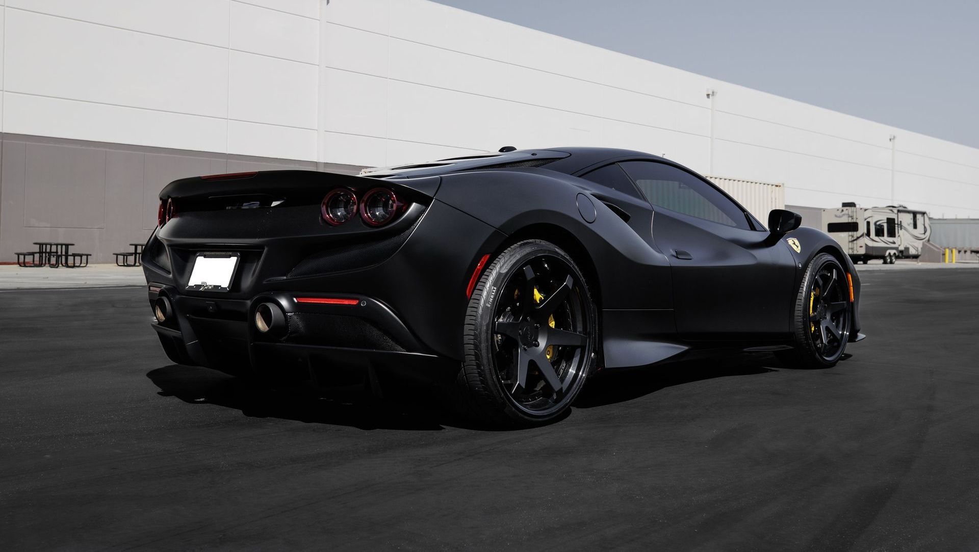 Black sports car parked on asphalt in front of a plain white wall, rear three-quarter view