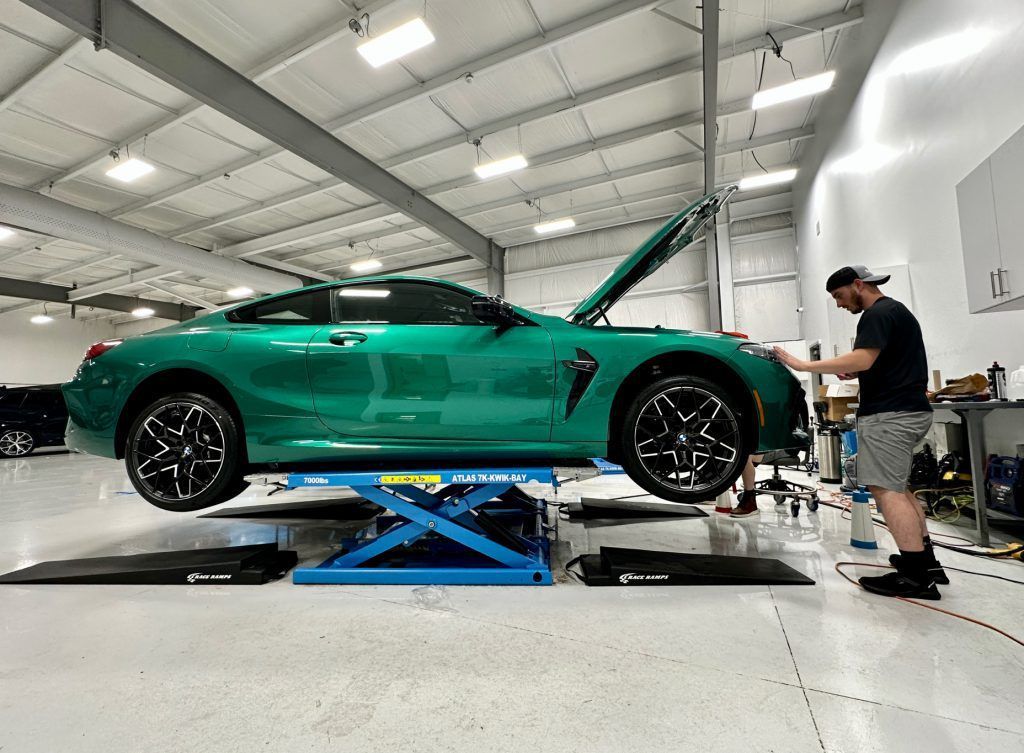 A man is working on a green car on a lift in a garage.