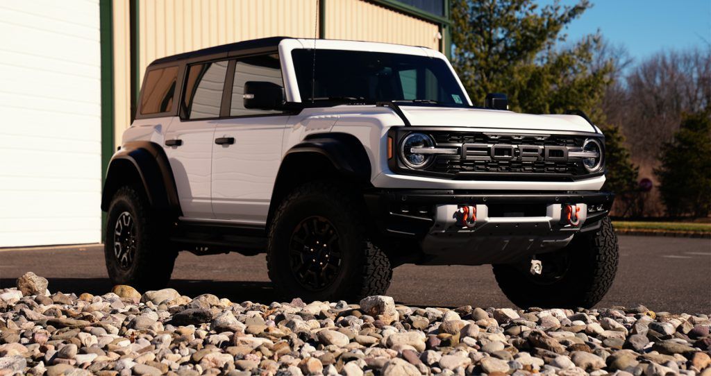 A white ford bronco raptor is parked on rocks in front of a garage.