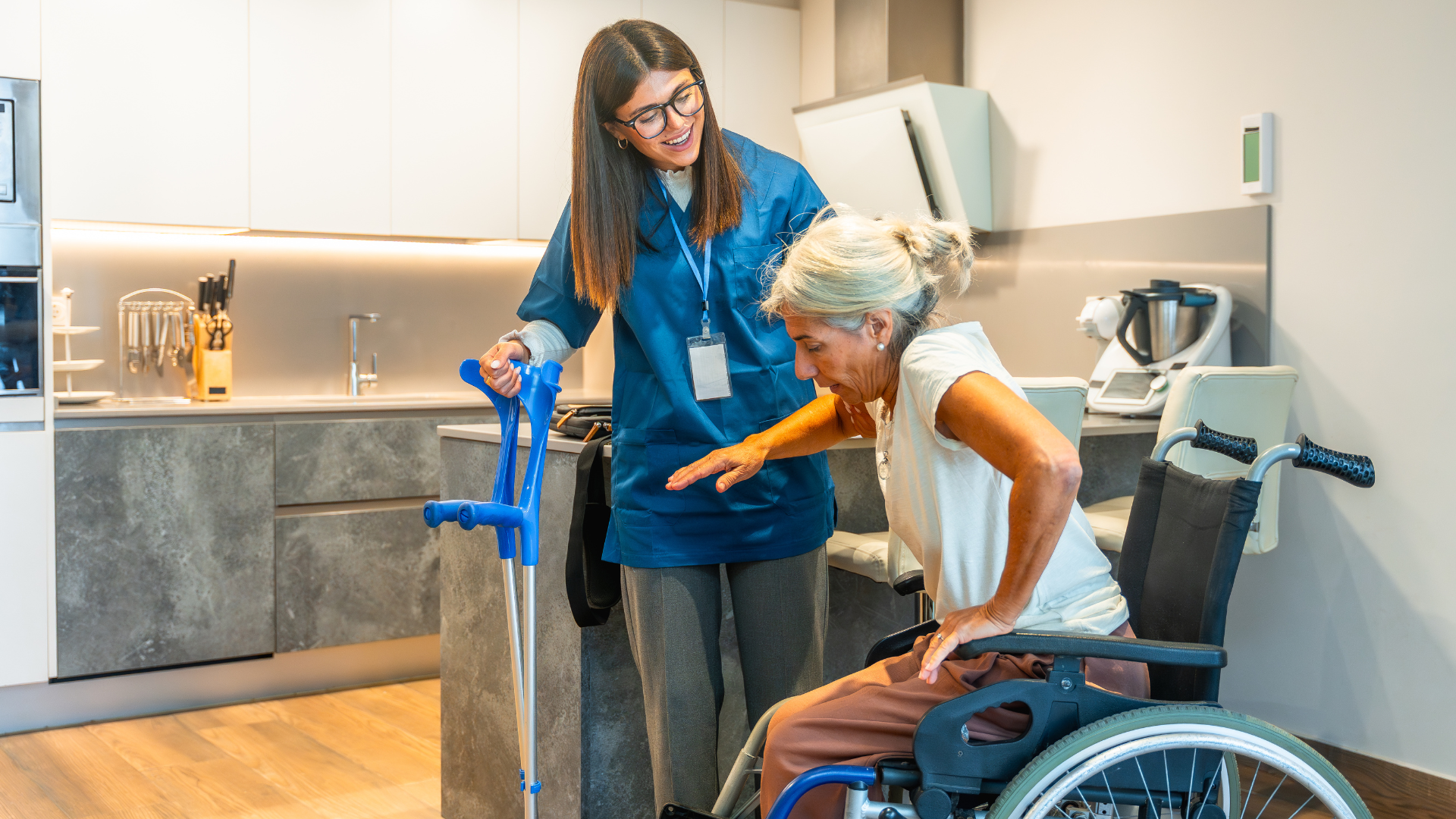 Caregiver assisting a person in a wheelchair, with crutches nearby, in a kitchen.