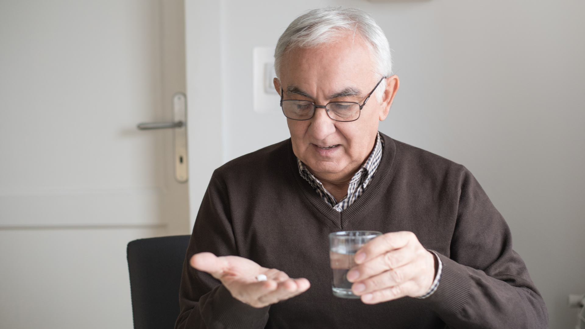 Older person in glasses holding pill in palm, drinking water.