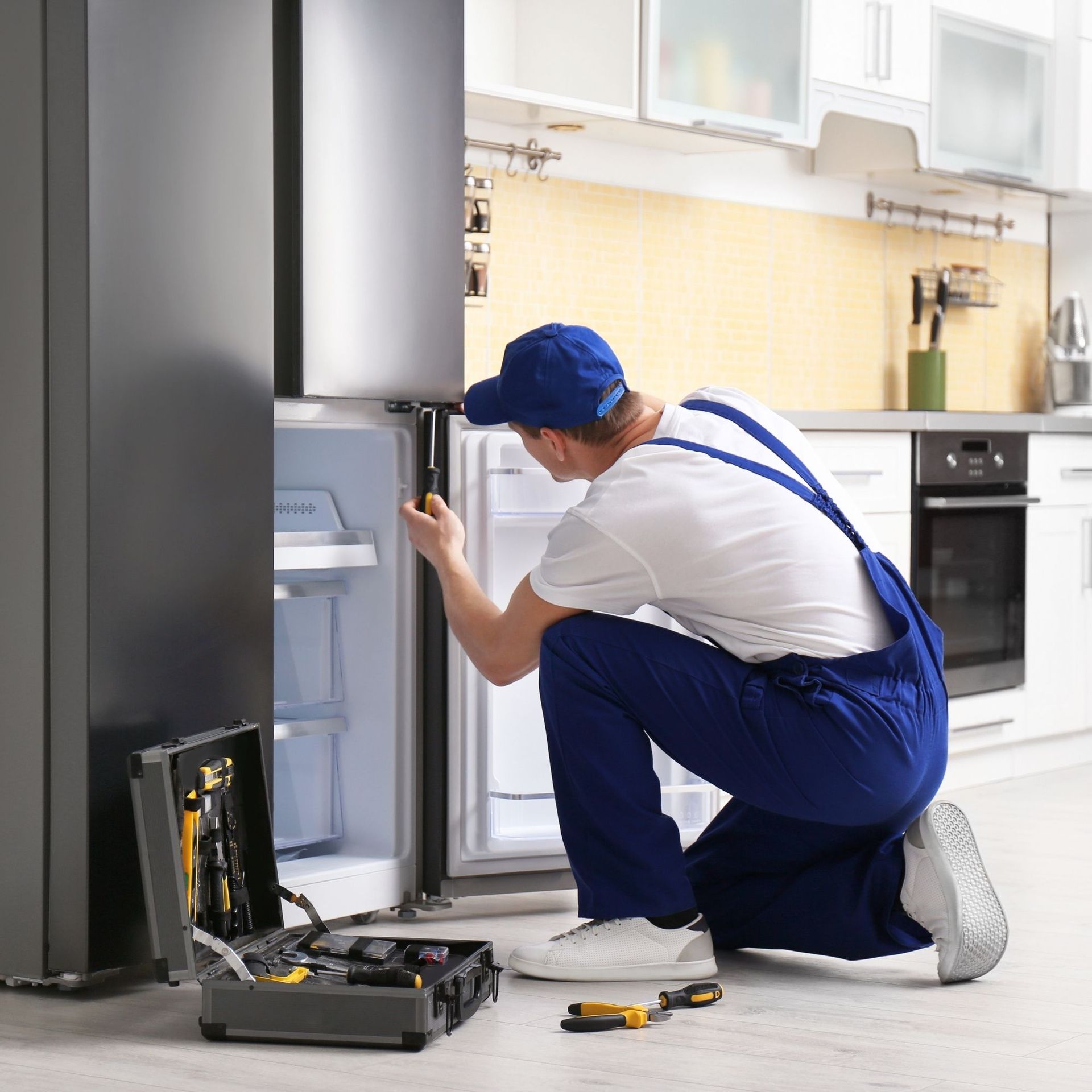 Skal East Inc - A repairman in blue overalls kneels in a kitchen, fixing a refrigerator door. A toolbox is open on the floor.