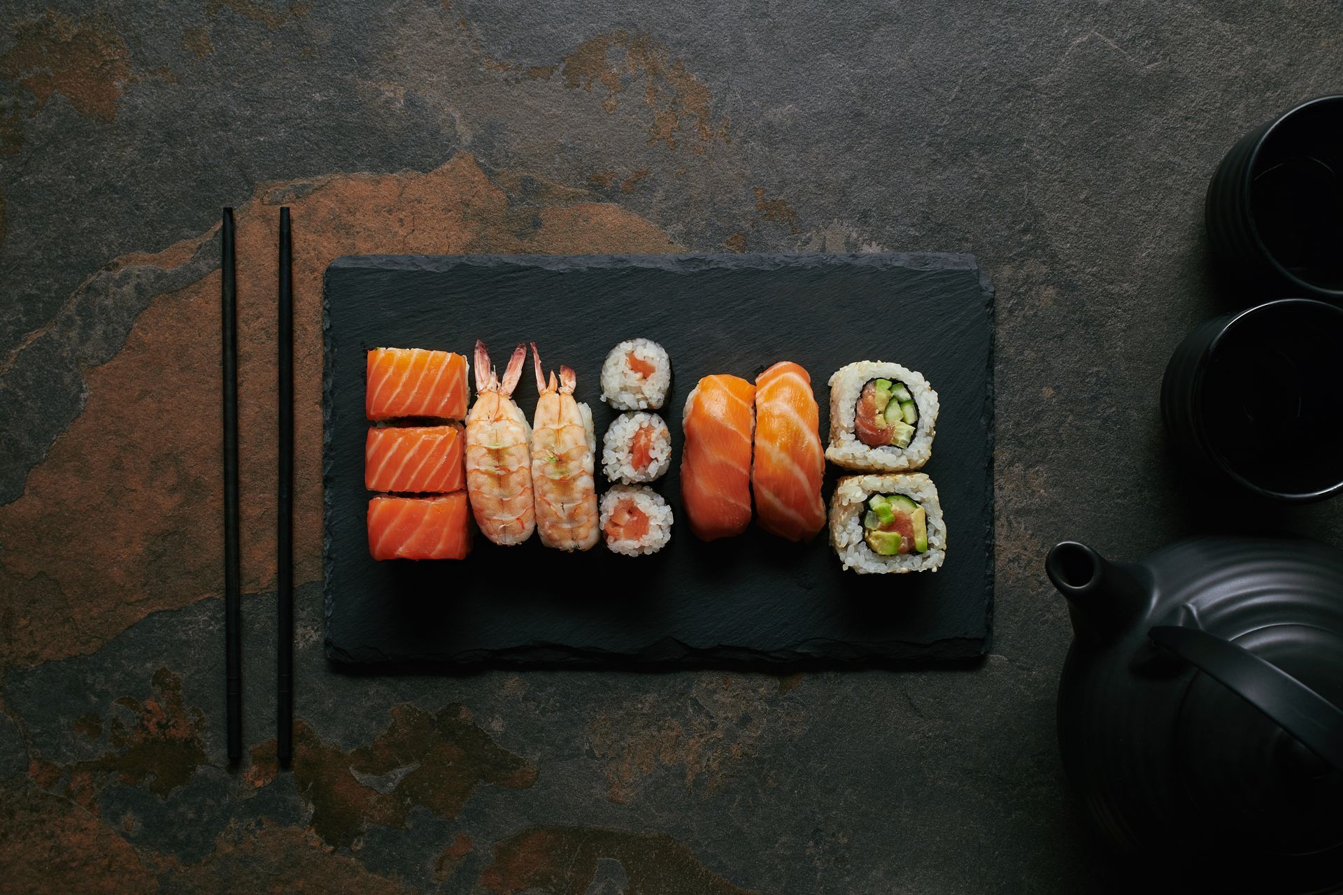 Sushi-Platte auf einer schwarzen Schieferplatte mit Essstäbchen und Teekanne auf einem dunkel strukturierten Tisch.