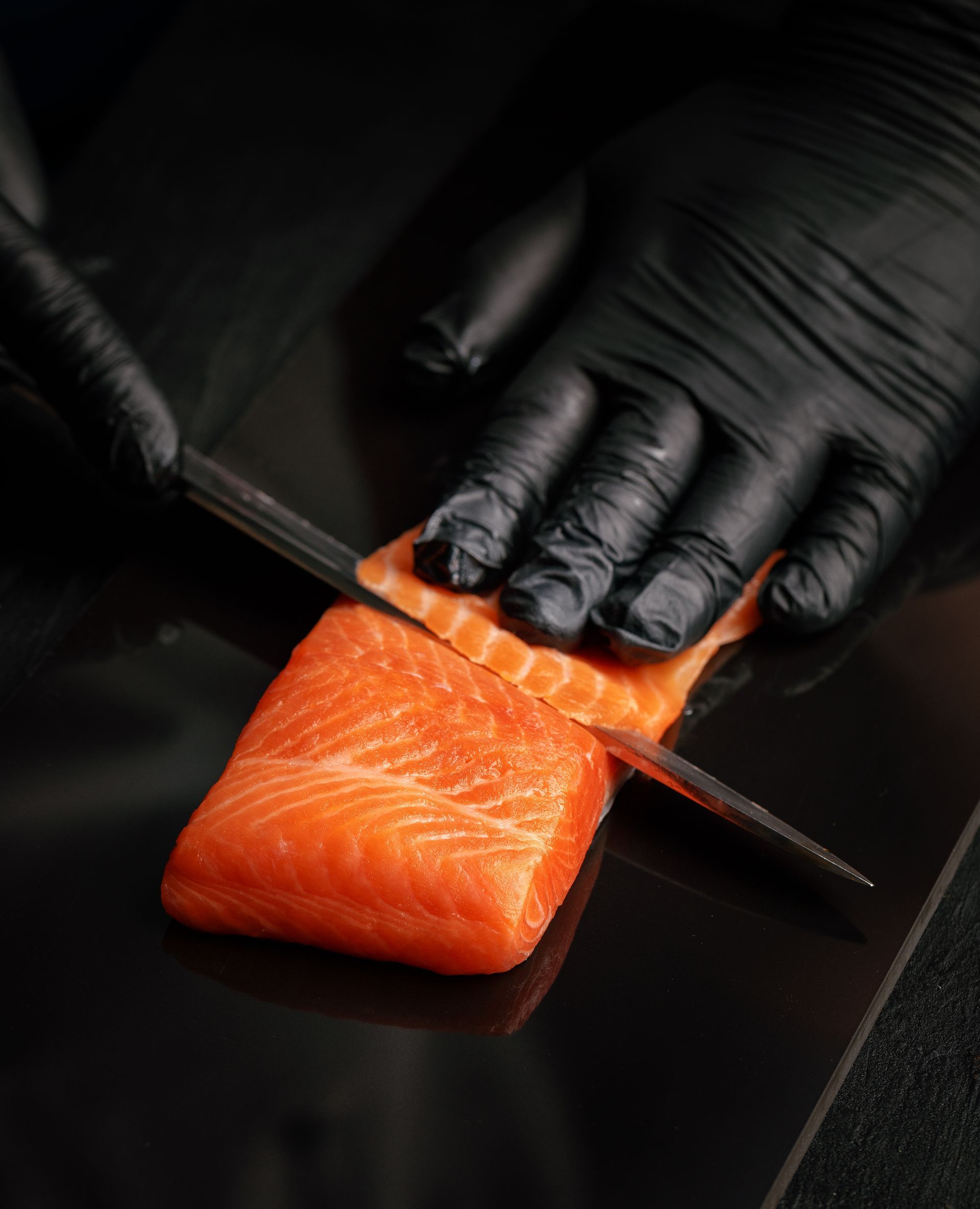 Behandschuhte Hände schneiden mit einem Messer ein orangefarbenes Lachsfilet auf einem schwarzen Schneidebrett