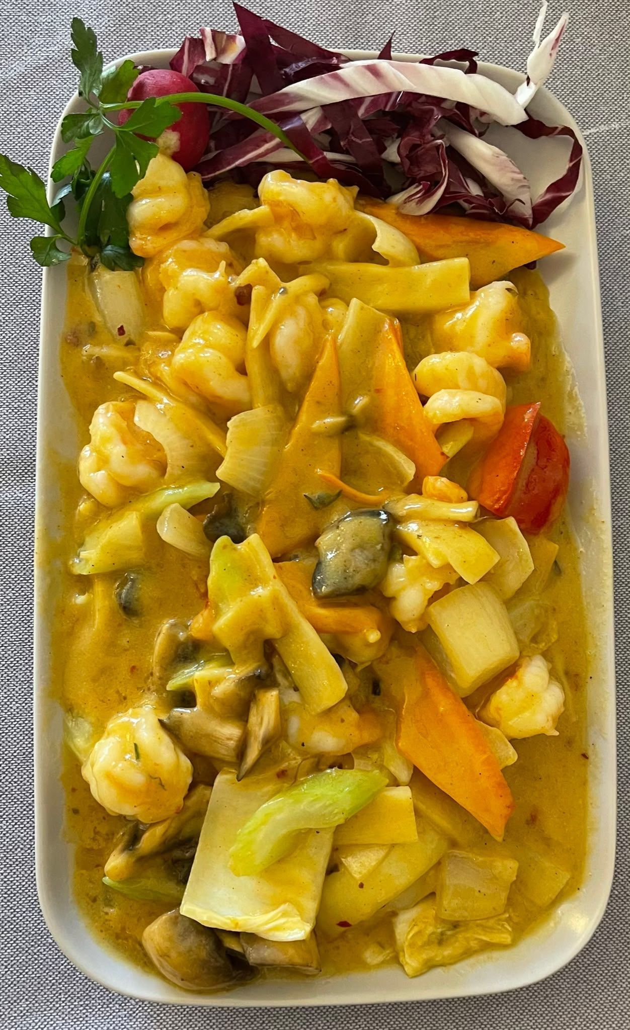 Weißer Teller mit gelbem Curry und Gemüse, garniert mit roten Blattgemüsen, auf einer strukturierten Tischmatte.