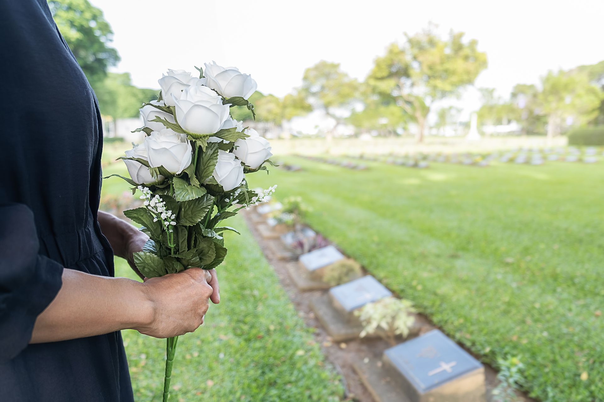 Person holding a bouquet of white roses in a grassy cemetery with graves and trees in the background