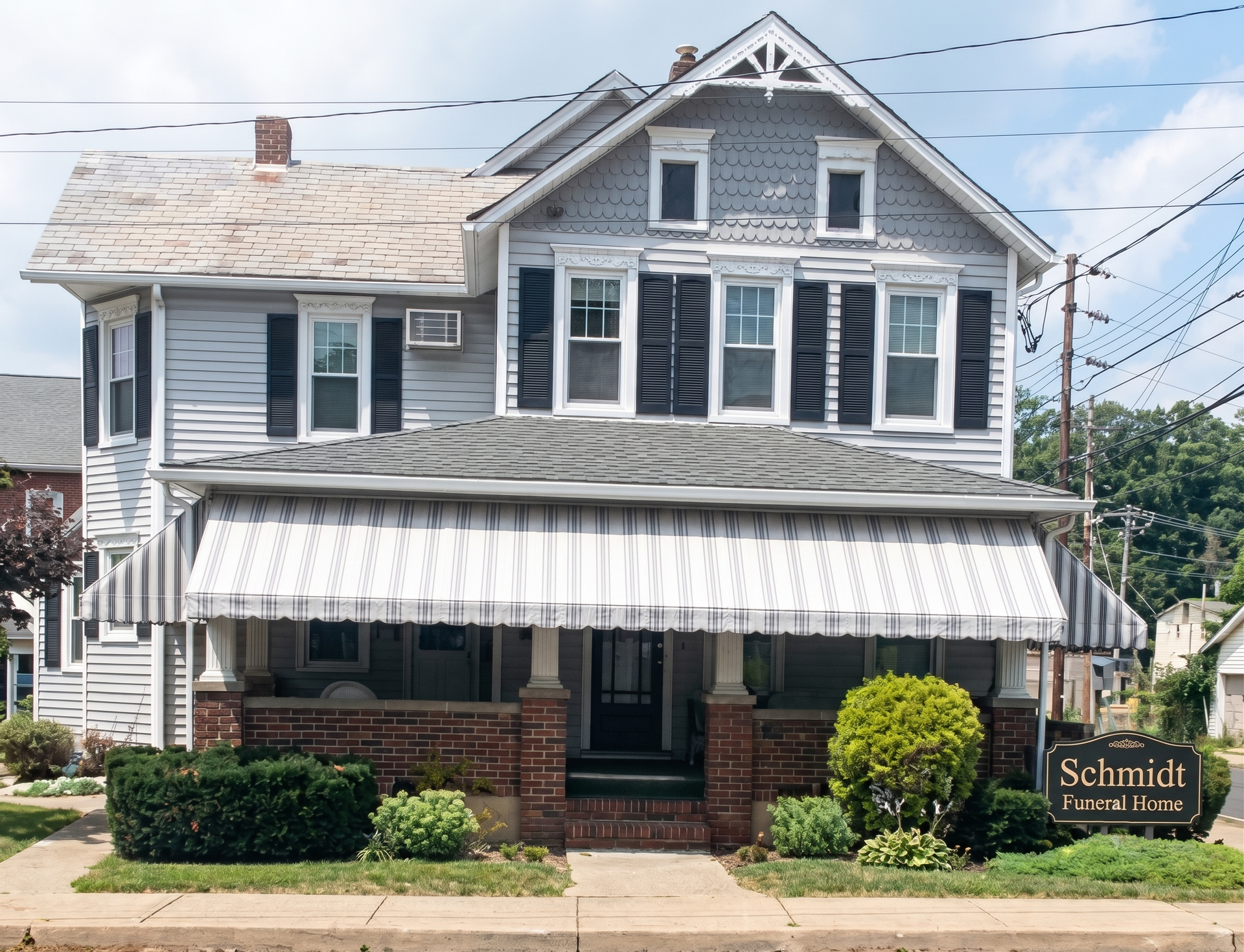 Two-story gray house with white trim, front porch, striped awning, and manicured shrubs by the sidewalk