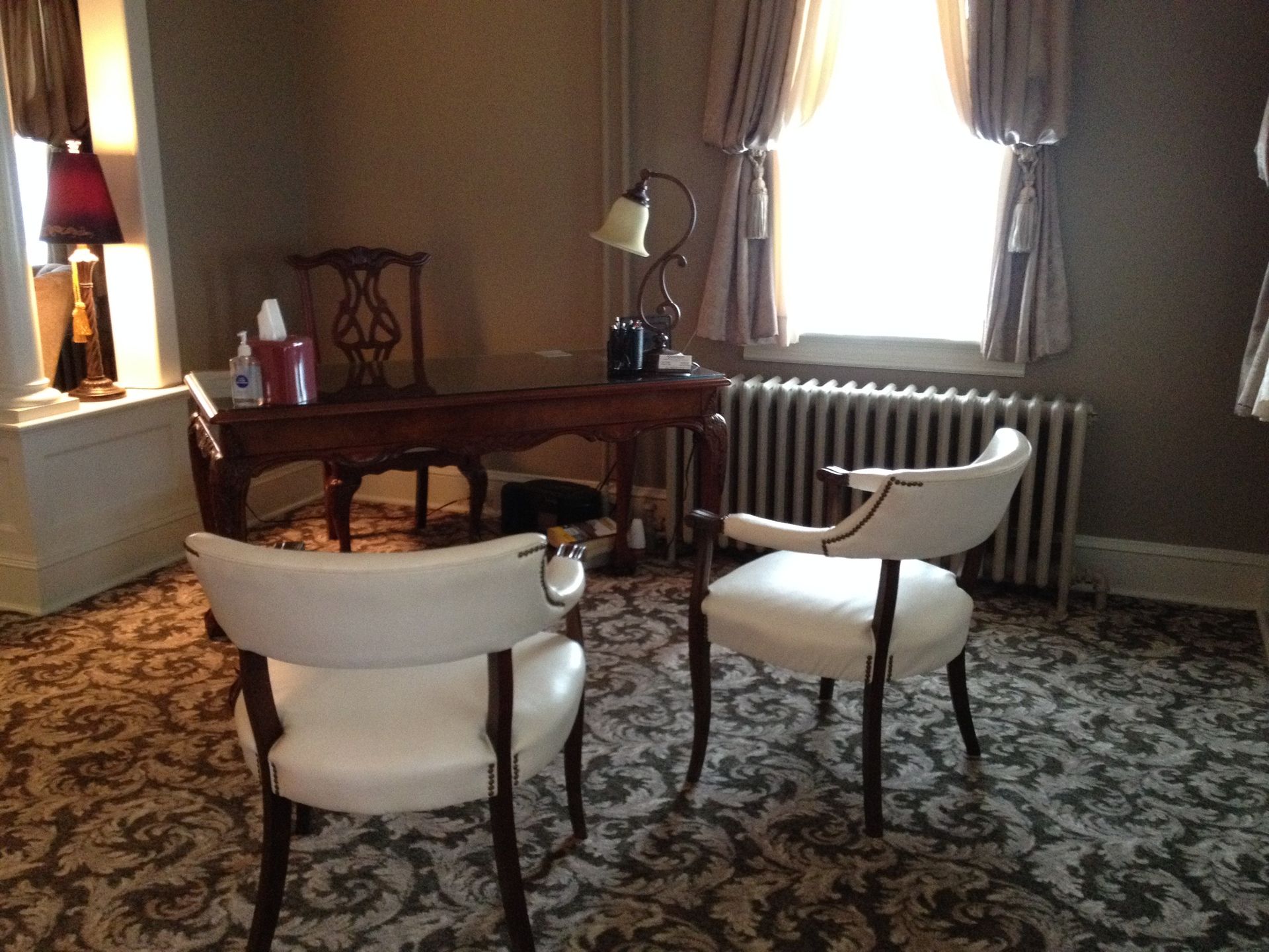Elegant living room with two white chairs, wooden desk, lamp, and bright window beside a radiator
