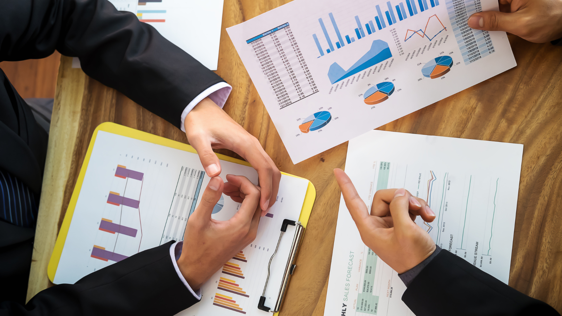 Hands pointing at financial charts on a wooden table.