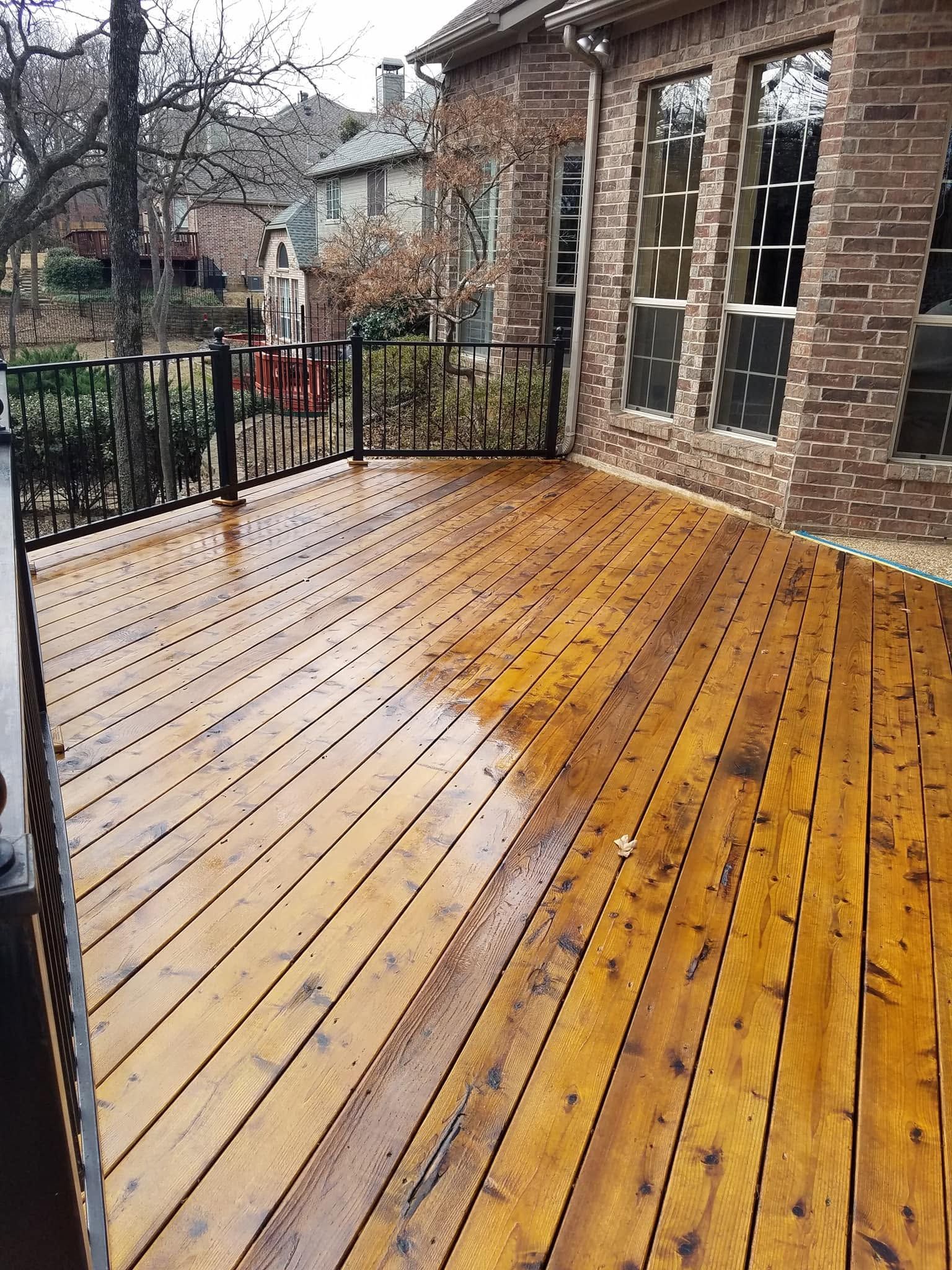 Rain-soaked wooden deck beside brick house and black railing, with yard and trees beyond