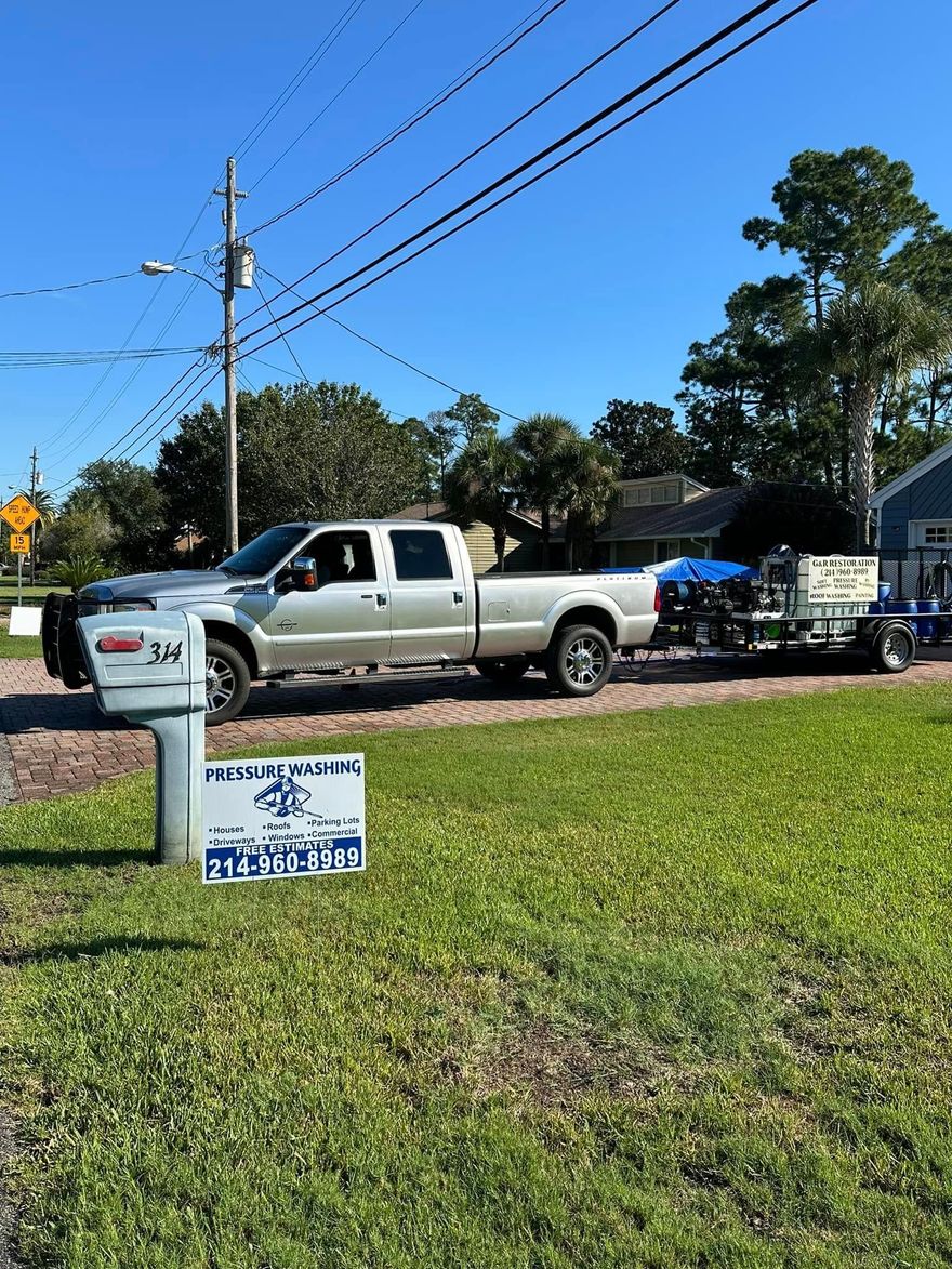 White pickup truck towing a trailer on a roadside beside a grassy yard and utility poles under a clear blue sky