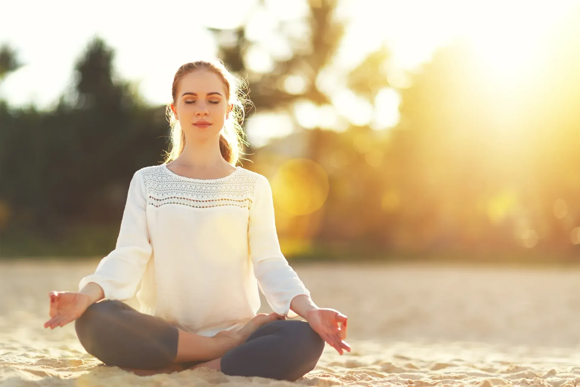 A woman is sitting in a lotus position on the beach with her eyes closed.
