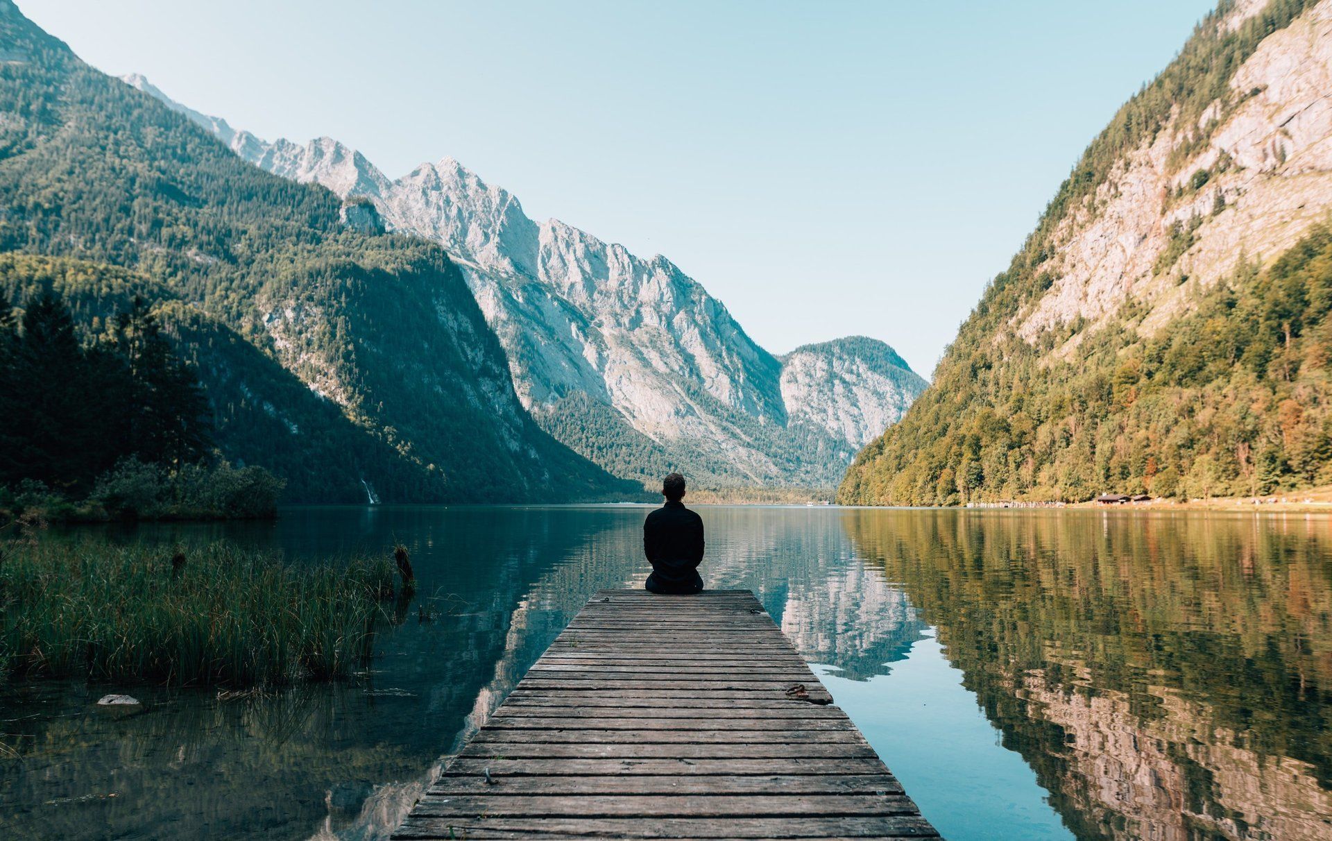 A man is sitting on a dock overlooking a lake with mountains in the background.