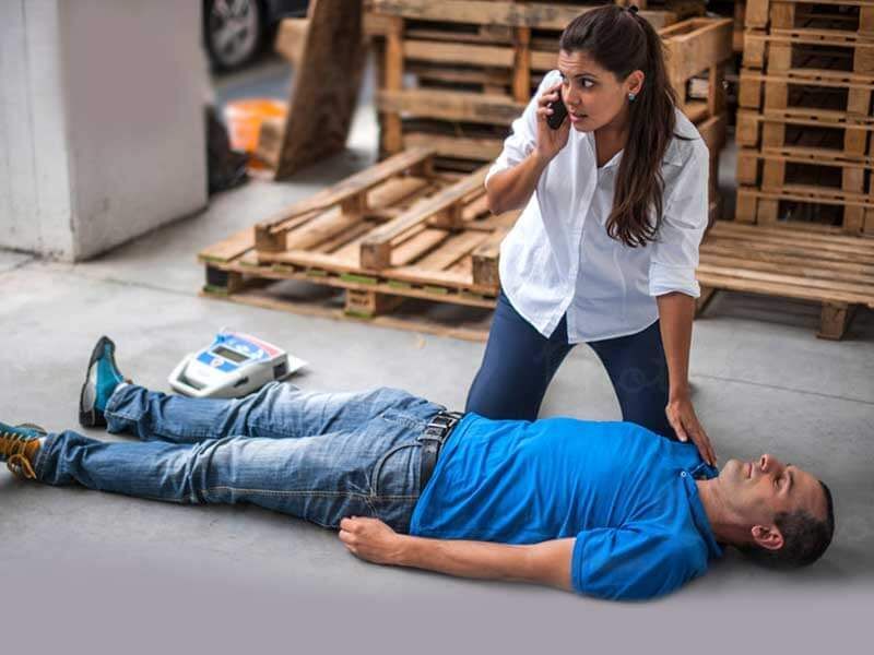 A person kneels beside an unconscious individual on the floor, calling for emergency help near wooden shipping pallets.