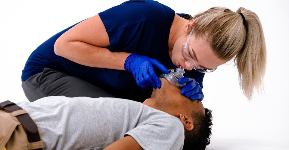 A responder in blue scrubs and gloves holds a clear resuscitation mask over a person’s mouth and nose to provide oxygen.