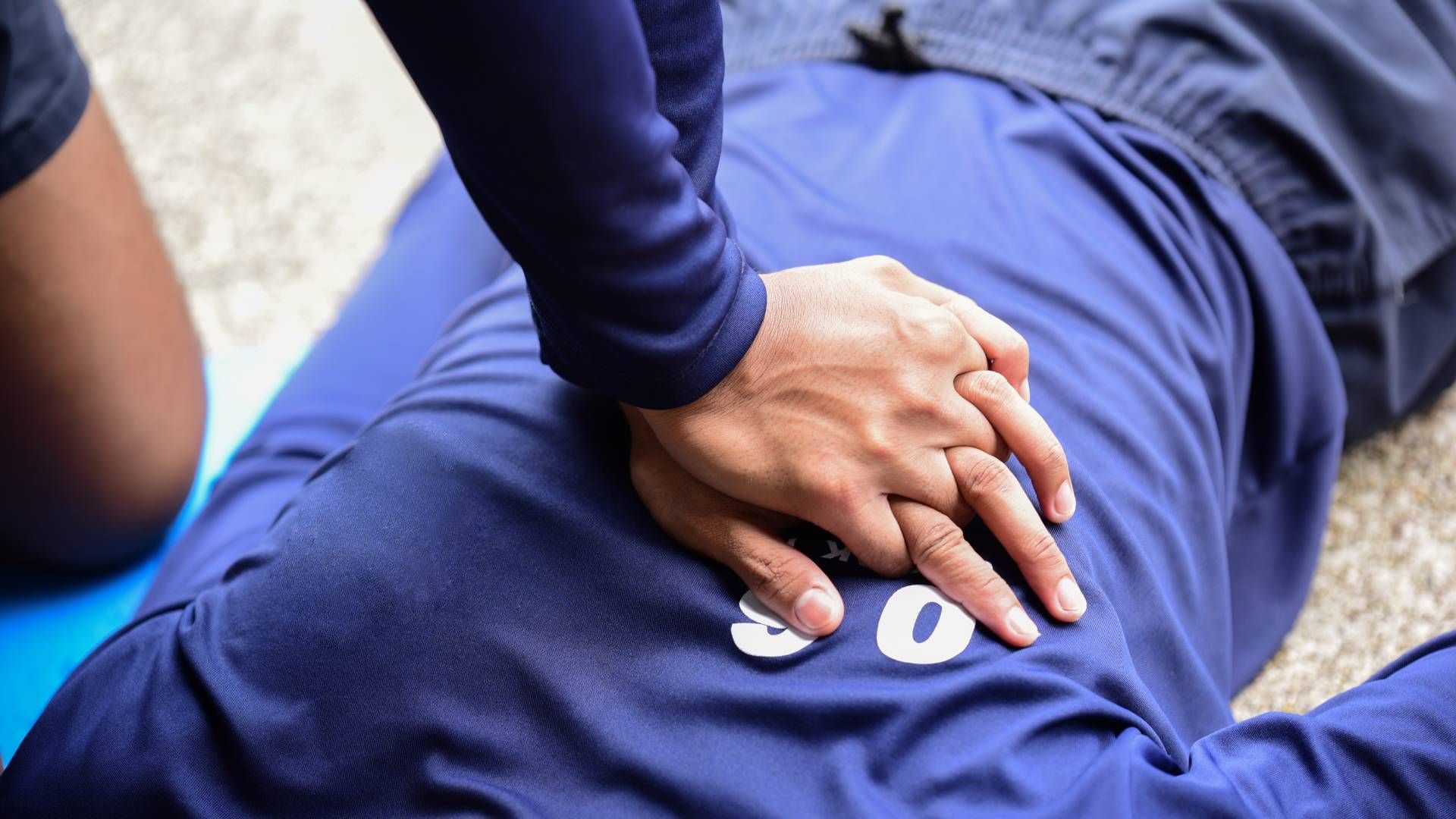 Hands are shown performing chest compressions on an individual in a blue shirt during CPR training.