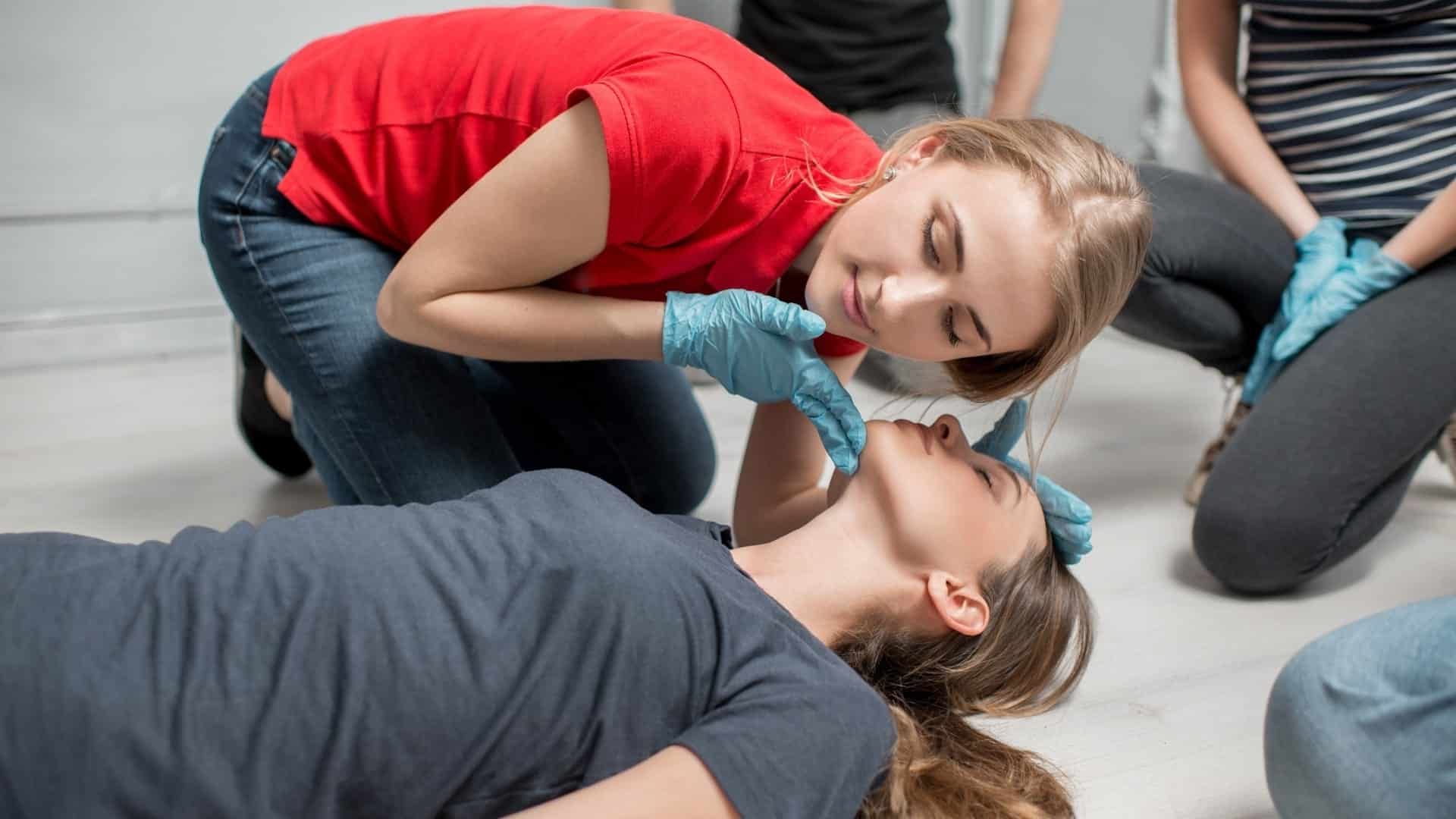 A person in a red shirt wears blue gloves and tilts the head of an unconscious person back to open their airway.