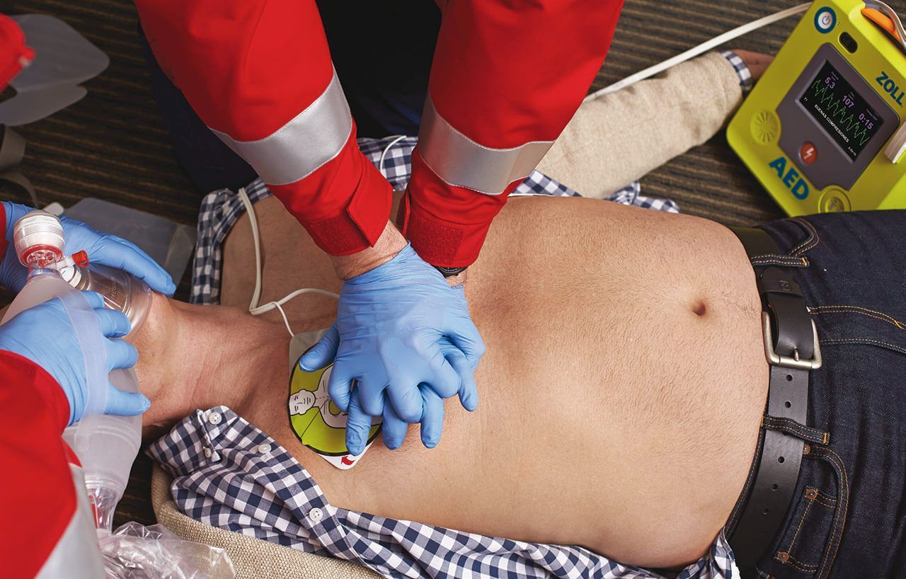 Medical personnel performing CPR on a patient, with an AED attached to the chest and a mask over their nose and mouth.