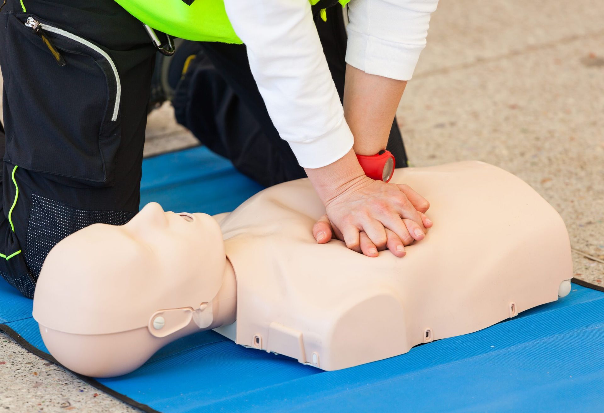 A person in a high-visibility vest performs CPR chest compressions on a medical training mannequin on a blue mat.