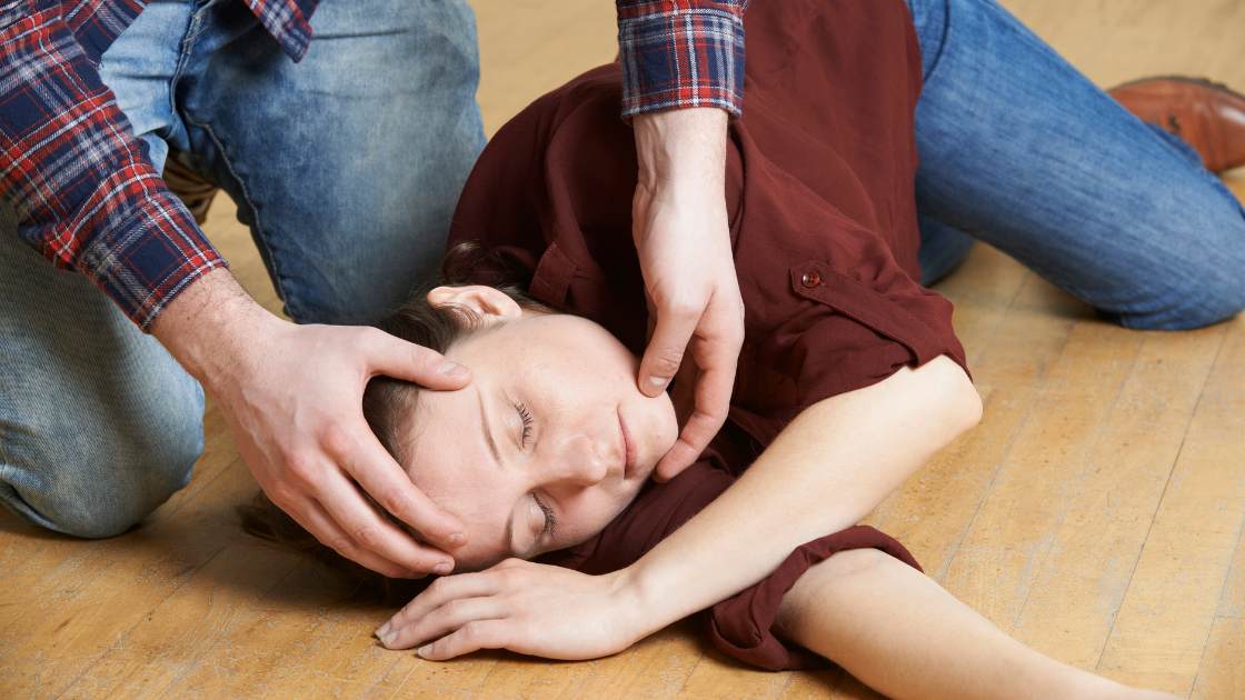 A person kneeling on a floor supports the head of another person who is lying down with eyes closed.
