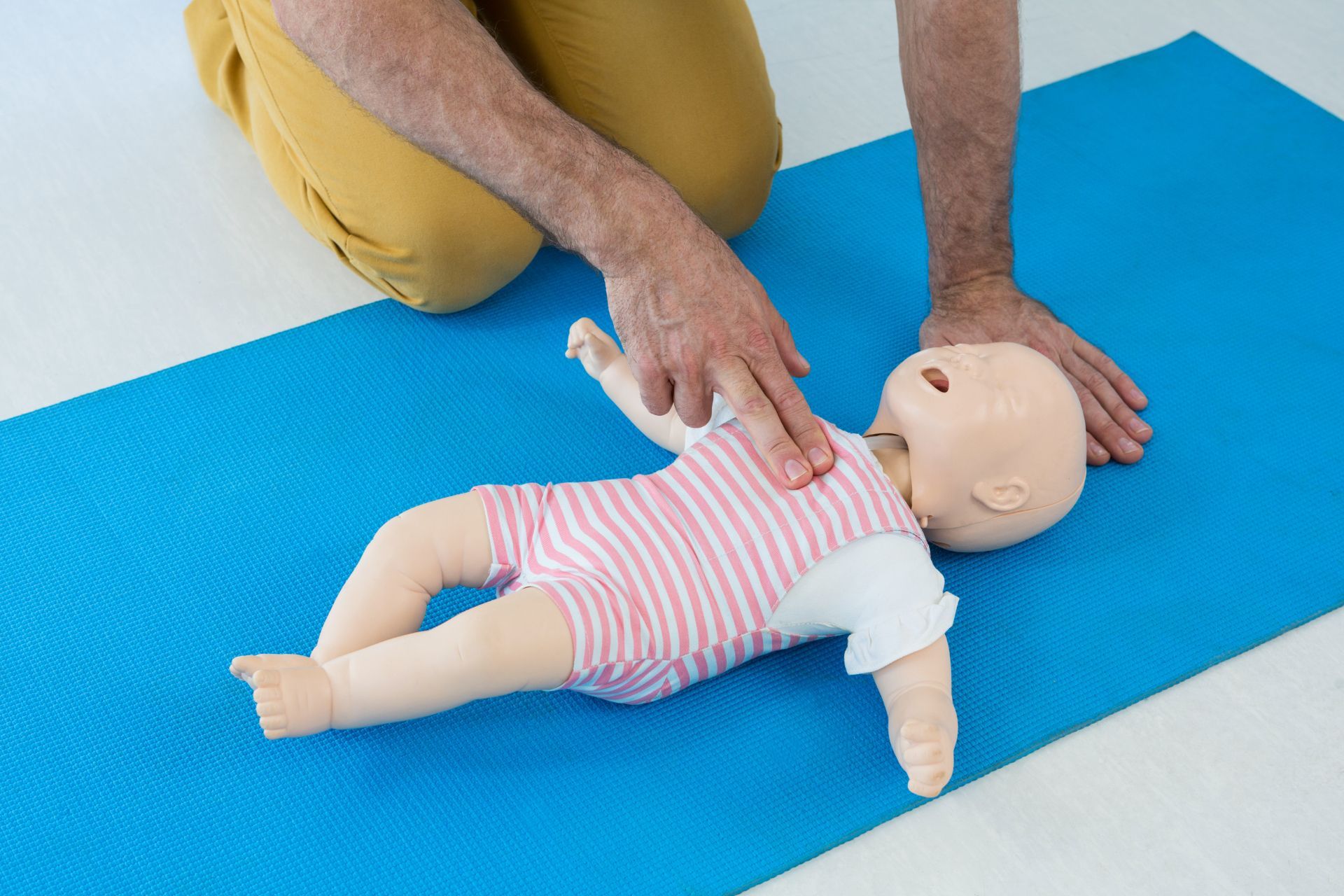A person performs CPR chest compressions on an infant medical mannequin lying on a blue yoga mat.