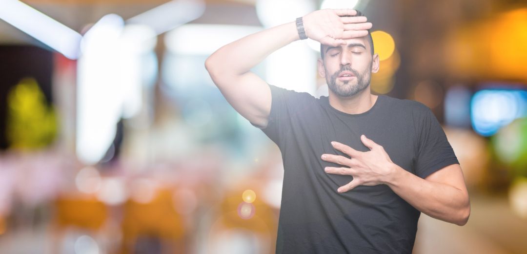 Man in black shirt shielding his eyes from bright sunlight, standing outdoors with blurred city lights behind him