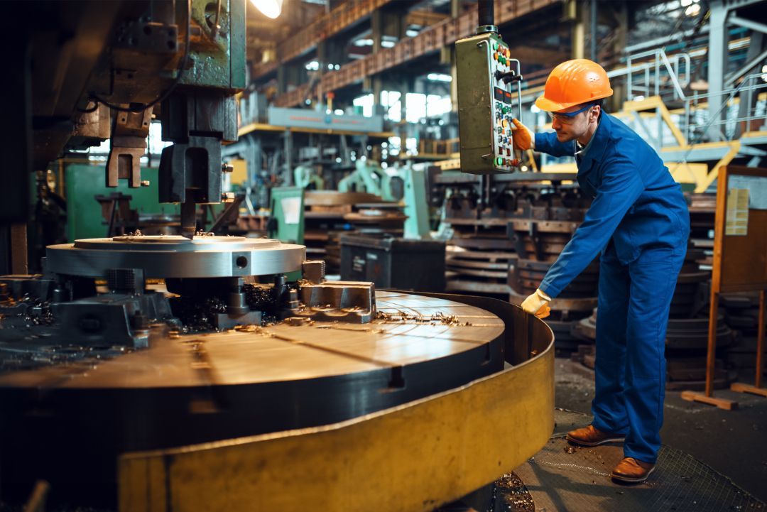 Worker in blue coveralls and orange hard hat operating machinery in an industrial factory.